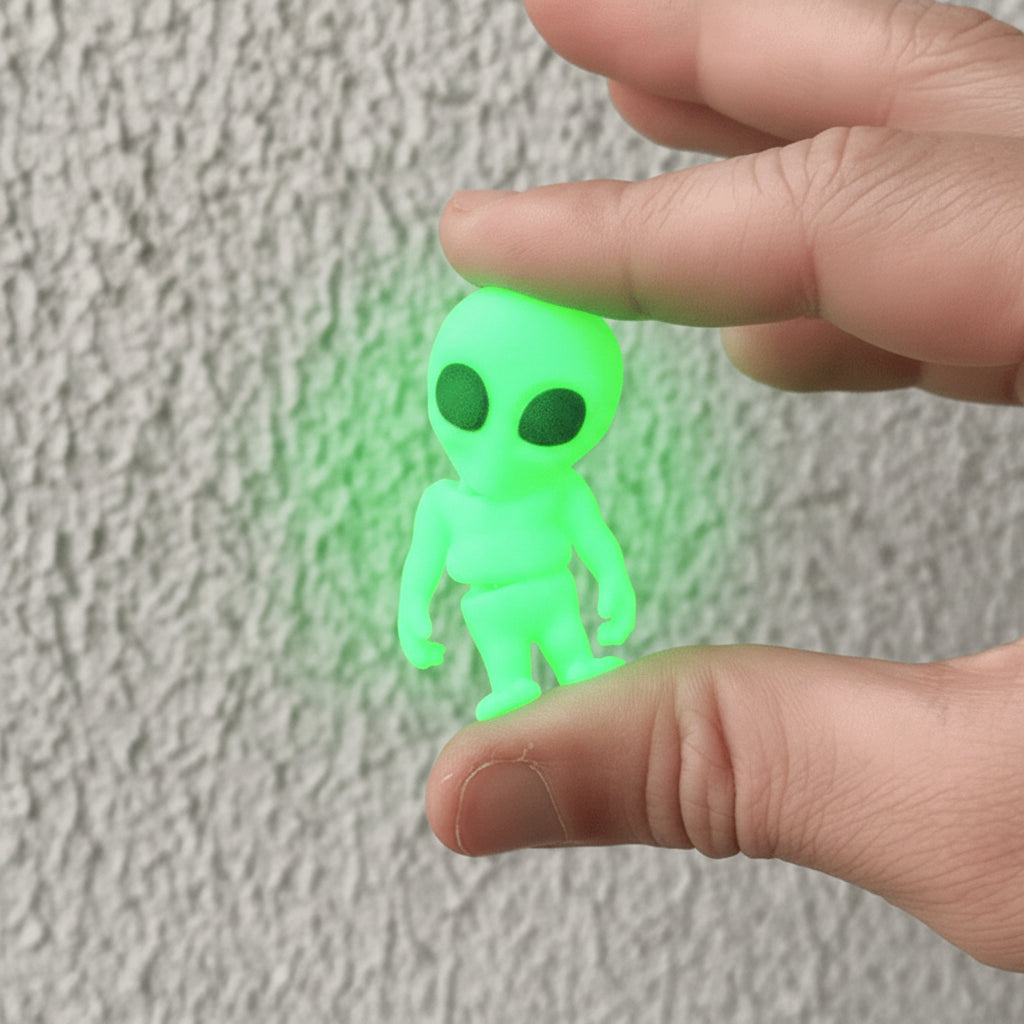 A small green alien figurine with large black eyes is held between a person's fingers against a textured light gray wall background.