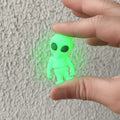 A small green alien figurine with large black eyes is held between a person's fingers against a textured light gray wall background.